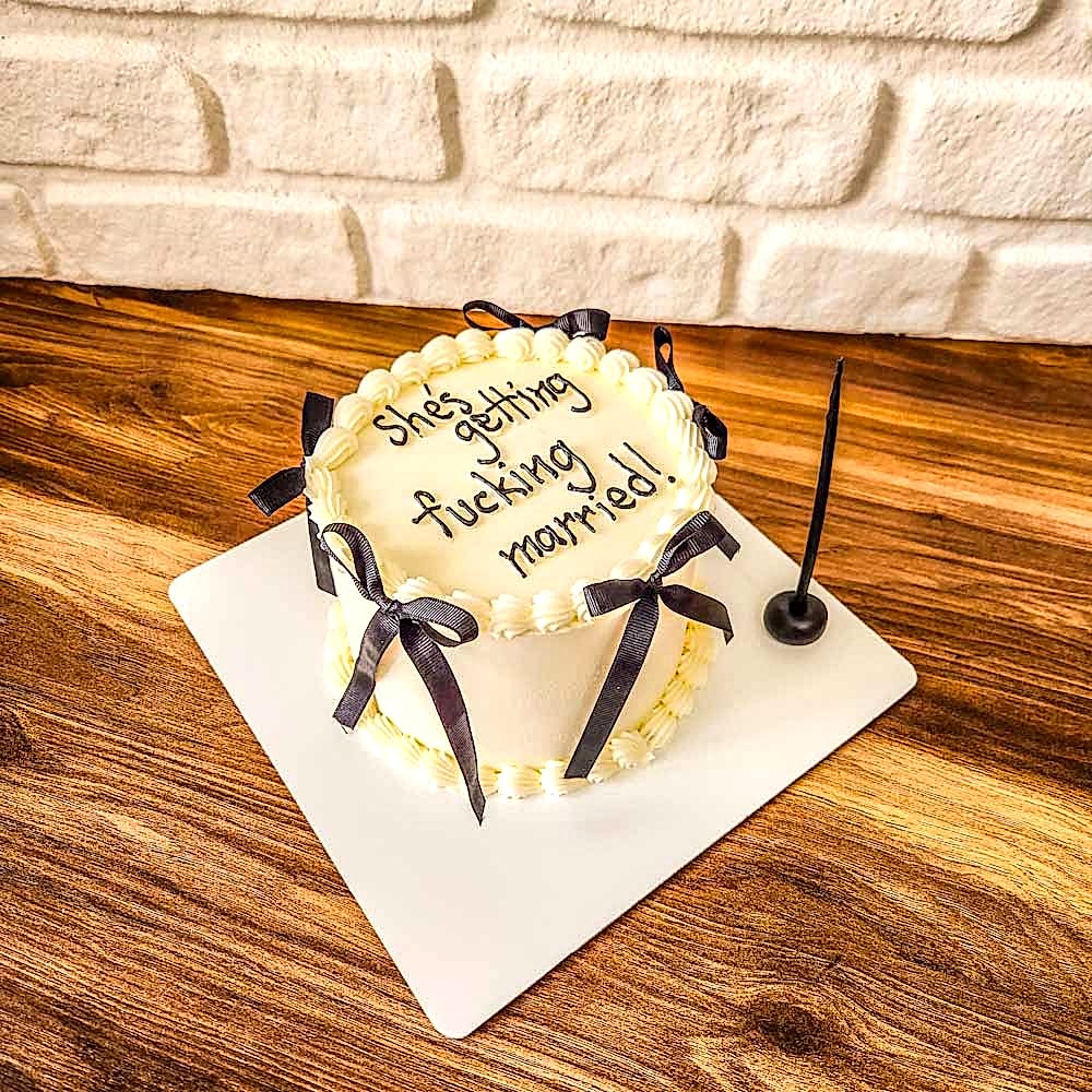 Beyaz zeminli butik pasta, üzerinde “She’s Getting Married!” yazısı ve zarif detaylarla süslenmiş; nişan, bridal shower veya bekarlığa veda kutlamaları için özel tasarım.
