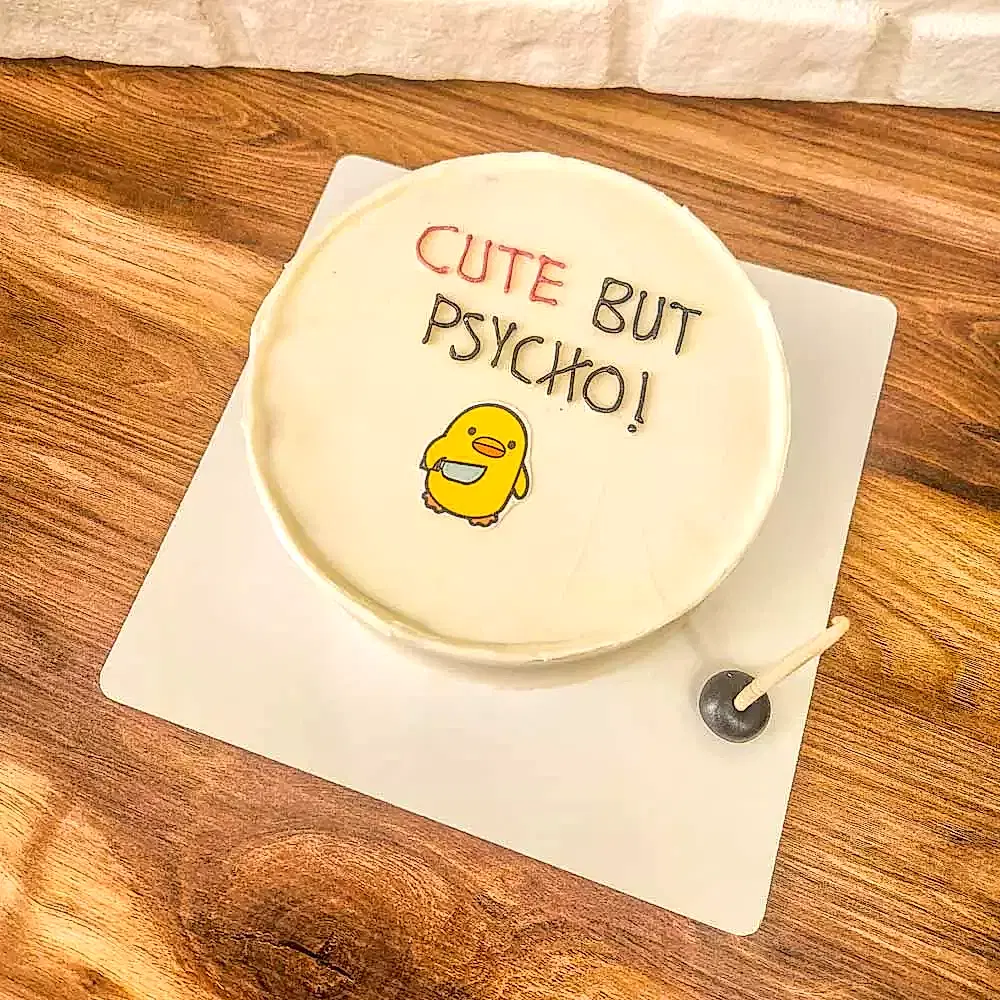 Beyaz zeminli butik pasta, üzerinde “Cute But…” yazısı ve elinde minik bıçak tutan civciv figürüyle süslenmiş; sevimli ama asi ruh halini mizahi bir dille yansıtan eğlenceli tasarım.