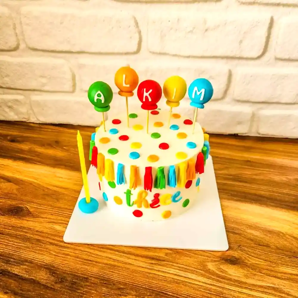 Renkli ponponlar, balon harfler ve “three” yazısıyla süslenmiş neşeli bir doğum günü pastası; Alkım’ın 3. yaş kutlamasına özel, enerjik ve çocuk ruhunu yansıtan bir tasarım.